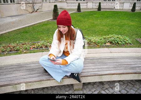 Young redhead girl student, sits on bench and uses smartphone app, watches videos online, sends message on mobile phone Stock Photo