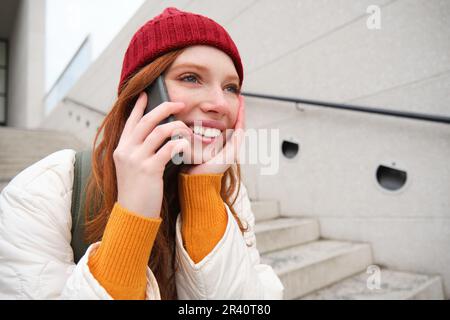 Beautiful smiling redhead female model, sits on street and talks on mobile phone, uses smartphone app to call abroad, laughing d Stock Photo