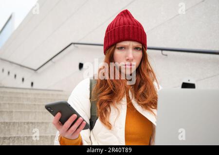 Portrait of redhead woman sits on stairs, uses laptop and holds smartphone, looks confused and upset at computer screen, tries t Stock Photo