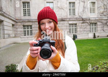 Redhead girl photographer takes photos on professional camera outdoors, captures streetstyle shots, looks excited while taking p Stock Photo