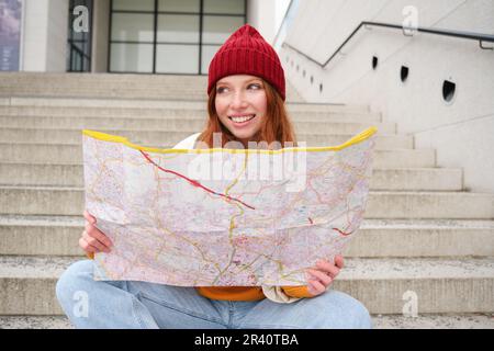 Young smiling redhead girl, tourist sits on stairs outdoors with city paper map, looking for direction, traveller backpacker exp Stock Photo