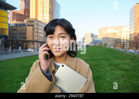 Stylish smiling girl, university student in trench coat, holds tablet, talks on mobile phone, has conversation over telephone an Stock Photo