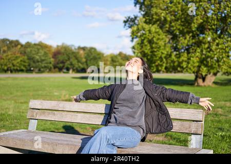 Portrait of young woman in outdoor clothes, sitting on bench relaxed, smiling and enjoying view on green park Stock Photo