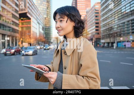 Woman walking on street using digital tablet, holding gadget in hands and looking away, standing in city centre Stock Photo