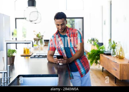Happy biracial man leaning on countertop and using smartphone in kitchen Stock Photo