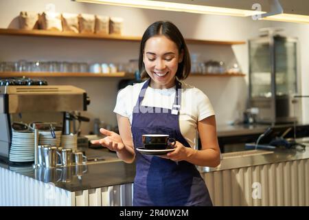 Portrait of smiling asian female barista, making coffee, holding cup of tea and taking it to cafe client, wearing apron, standin Stock Photo