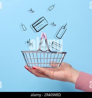 A woman's hand holds an empty shopping cart and icons of cards, coupons on a blue background. Sale and discount concept Stock Photo
