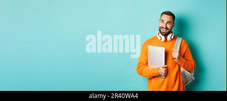 Happy man with backpack and headphones, holding laptop and smiling, looking left thoughtful, standing over turquoise background Stock Photo