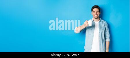 Smiling bearded man holding mug and drinking coffee, standing against blue background Stock Photo