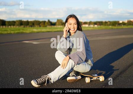 Cute teen girl sits on skateboard and talks on mobile phone. Happy skater woman having conversation on smartphone Stock Photo