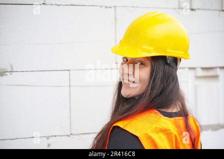 Young woman in yellow hard hat and orange high visibility vest, long dark hair, hands crossed, looking over her shoulder, smiling confident. Blurred c Stock Photo