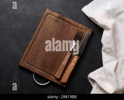 Empty wooden kitchen cutting board, knife and fork with wooden handle on black background, top view Stock Photo
