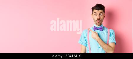 Intrigued young man pointing finger left at logo, checking out promo offer, standing in bow-tie and classy outfit, pink backgrou Stock Photo