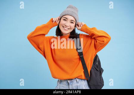 Cute smiling asian girl puts on warm hat to go outside, walks with backpack in orange sweater, blue background Stock Photo