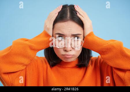 Frustrated young woman in glasses, holds hands on head complicated, troubled by problems, stands over blue background Stock Photo
