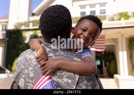 Happy african american boy with flag of america embracing army soldier father outside house Stock Photo