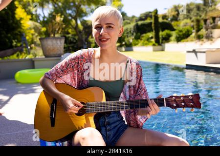 Portrait of happy caucasian woman having pool party, playing guitar and smiling in garden Stock Photo