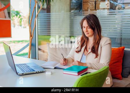 Portrait of young businesswoman working in office using laptop. Middle-aged woman writing down notes in notebook Stock Photo