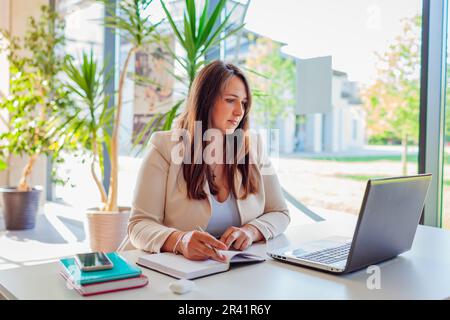 Young female teacher getting ready for lesson working in office using laptop. Woman writing notes in notebook Stock Photo