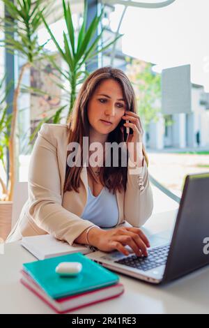 Female office worker talking on smartphone using laptop. Young businesswoman working online through modern technologies Stock Photo