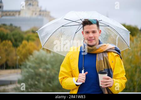 Young man wearing a yellow raincoat with a coffee and umbrella in street in autumn. Stock Photo