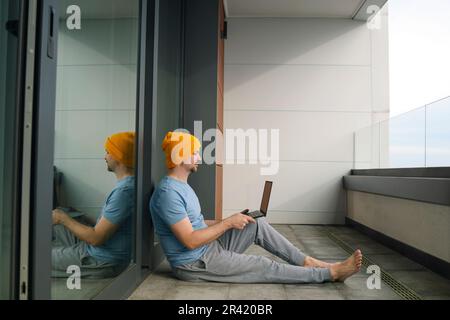 Young man in casual clothes and barefoot sits on balcony with laptop. Male freelancer in yellow hat, blue t-shirt and gray pants Stock Photo