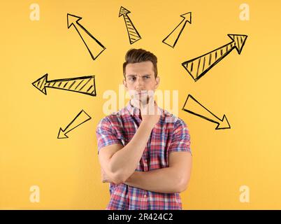 Choice in profession or other areas of life, concept. Making decision, thoughtful young man surrounded by drawn arrows on yellow background Stock Photo
