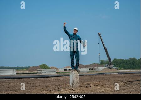 Builder in a protective helmet holds a laptop and stands on a pile in the middle of a construction site.  Stock Photo