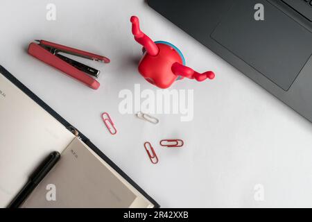 Flat lay design of work desk with notebook, pen, laptop, stapler, paper clips, and a toy on white background. Stock Photo