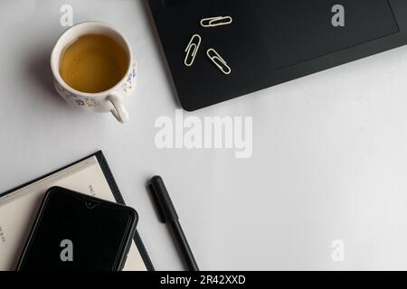 Flat lay design of work desk with notebook, pen, laptop, mobile, teacup, and paper clips on white background. Stock Photo
