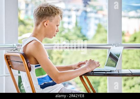 Preteen boy sitting on hotel balcony and surfing the internet on grey laptop. Schoolboy enjoys spending summer holidays and playing games Stock Photo