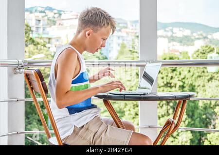 Preteen boy sitting on hotel balcony and surfing the internet on grey laptop. Schoolboy enjoys spending summer holidays and playing games Stock Photo