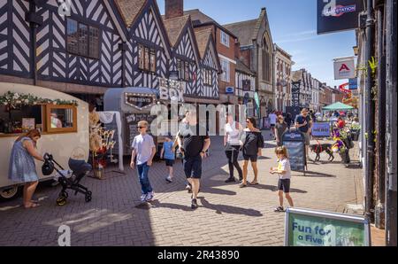 People enjoying the sun while walking along a busy street lined with shops and pop up food and drink stalls during Lichfield Food Festival. Stock Photo