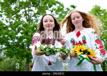 two beautiful young girls wearing embroidered shirts laughing smiling putting wreaths on the water facing the camera stretching out wreaths to us different sunflowers girl has red bright sunny hair Stock Photo
