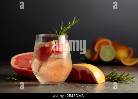 Cocktail gin tonic with ice, rosemary, and grapefruit on a stone table. In the background are various citrus fruits for making cocktails. Stock Photo