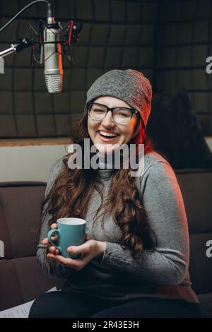Woman drinks hot tea or coffee from cup at cozy sound recording studio in the morning before broadcasting live. Beautiful female voice artist enjoying Stock Photo