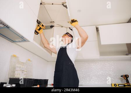 Maintenance man installing kitchen furniture in a black apron and yellow gloves using a power skrewdriver Stock Photo