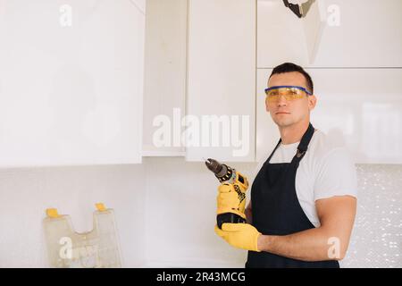 Maintenance man installing kitchen furniture in a black apron and yellow gloves using a power skrewdriver Stock Photo