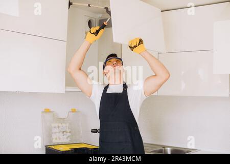 Maintenance man installing kitchen furniture in a black apron and yellow gloves using a power skrewdriver Stock Photo