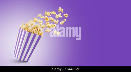 Popcorn flying out of purple white striped paper box isolated on purple background with copy space. Splash, levitation of popcorn grains. Stock Photo
