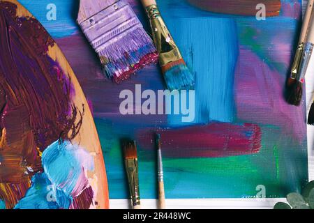 Canvas with colorful abstract painting, different brushes and wooden artist's palette on table, flat lay Stock Photo