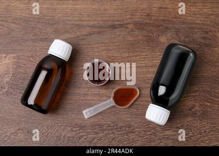 Bottles of syrup, measuring cup and dosing spoon on wooden table, flat lay. Cold medicine Stock Photo