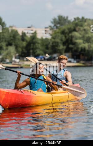 carefree interracial couple in life vests holding paddles and looking at clear water while sailing in sportive kayak during recreation weekend on summ Stock Photo