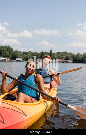 excited multiethnic couple in life vests spending summer weekend on picturesque lake while paddling in sportive kayak under blue sky with white clouds Stock Photo