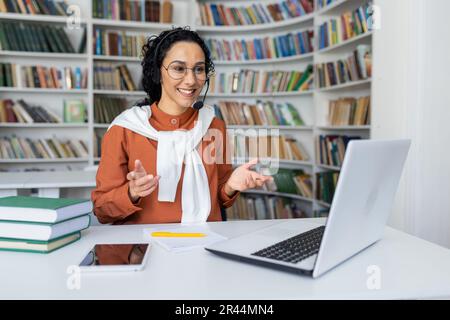 Teacher conducts lessons online, woman with video call headset smiling and explaining lecture online using laptop for remote communication and learning, sitting at desk in university library at desk. Stock Photo