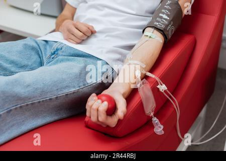 partial view of young man with transfusion set and blood pressure cuff sitting on ergonomic and comfortable chair and squeezing rubber ball in modern Stock Photo