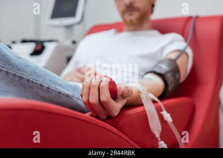 selective focus of rubber ball in hand of cropped volunteer with transfusion set sitting on comfortable medical chair near transfusion machine in bloo Stock Photo