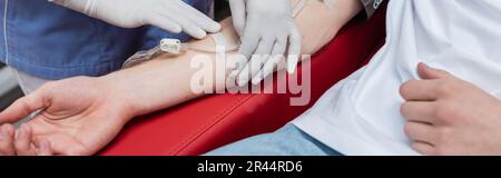 cropped view of multiracial nurse fixing blood transfusion set with band-aid on arm of young volunteer in contemporary blood donation center, banner Stock Photo