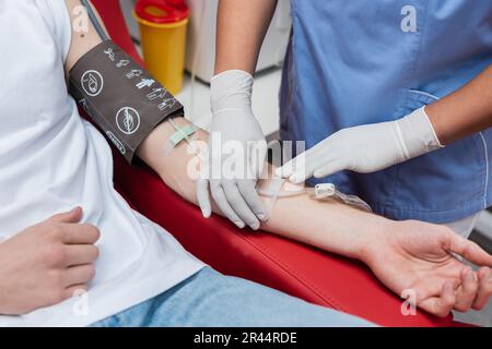 cropped view of multiracial healthcare worker in latex gloves sticking band-aid on arm of volunteer with blood pressure cuff and transfusion set in me Stock Photo