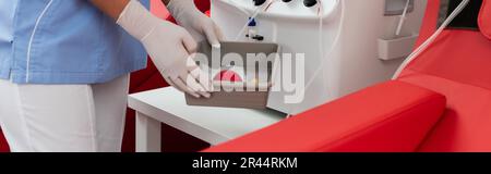 cropped view of multiracial nurse in uniform and latex gloves holding medical tray with rubber ball near transfusion machine in blood donation center, Stock Photo